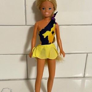 May include: A blonde doll wearing a yellow and black one-piece swimsuit with a yellow skirt. The doll has a long braid and is standing on a white tiled surface.