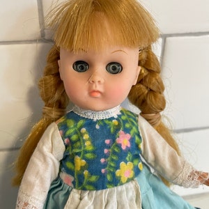 May include: A vintage doll with blonde hair in braids, wearing a blue and white floral dress with a white apron. The doll has large, green eyes and a painted smile.