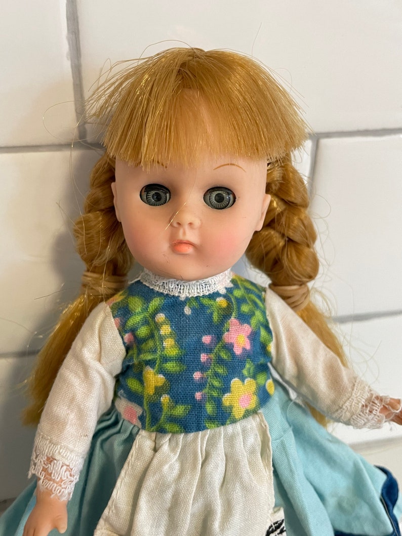 May include: A vintage doll with blonde hair in braids, wearing a blue and white floral dress with a white apron. The doll has large, green eyes and a painted smile.