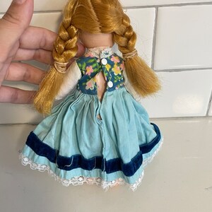 May include: A vintage doll with blonde hair in braids wearing a blue dress with a floral print bodice and a blue velvet trim. The doll has a white button closure on the back.