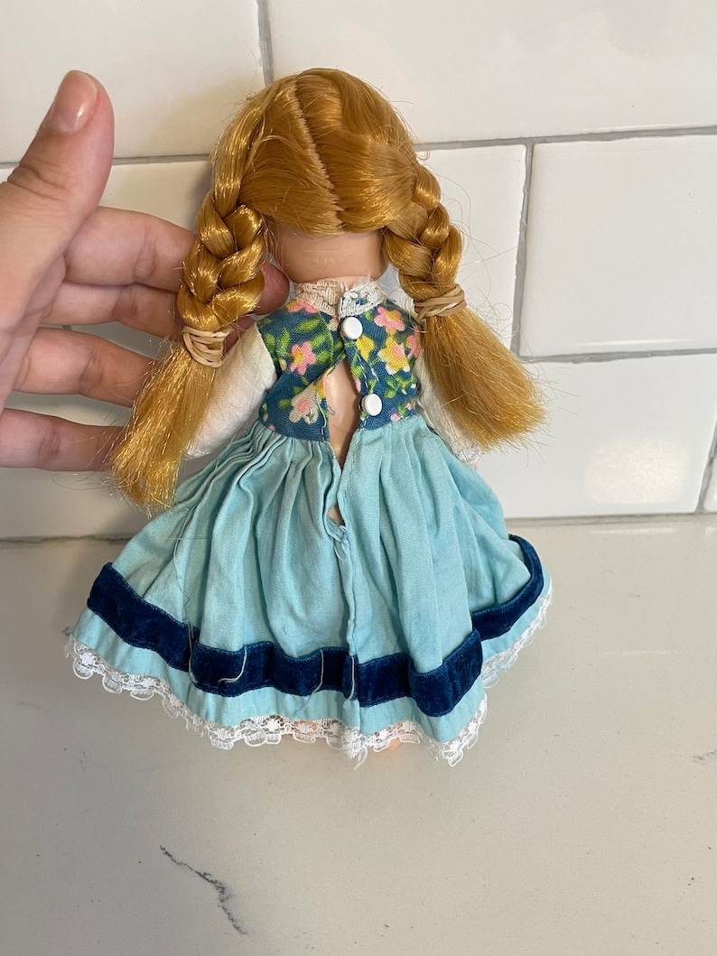 May include: A vintage doll with blonde hair in braids wearing a blue dress with a floral print bodice and a blue velvet trim. The doll has a white button closure on the back.