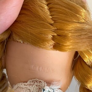 May include: A porcelain doll with blonde hair in a braid. The doll is wearing a blue and green plaid dress with a white lace collar and a pink flower print. The doll is marked "Ginny" on the back of the neck.