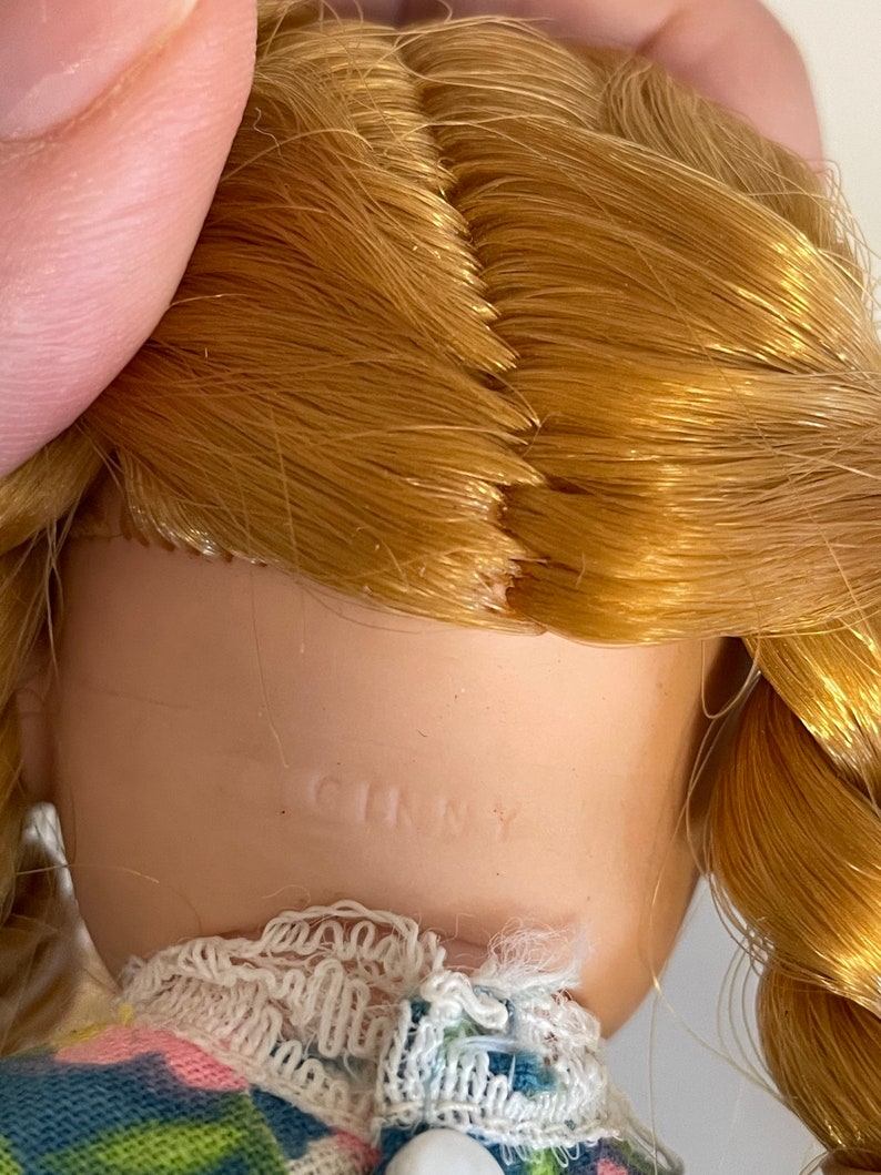 May include: A porcelain doll with blonde hair in a braid. The doll is wearing a blue and green plaid dress with a white lace collar and a pink flower print. The doll is marked "Ginny" on the back of the neck.