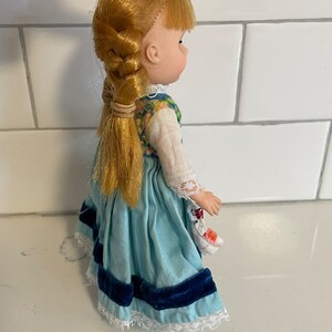 May include: A vintage porcelain doll with blonde hair in a braid, wearing a blue and white dress with a floral bodice and a blue velvet sash.