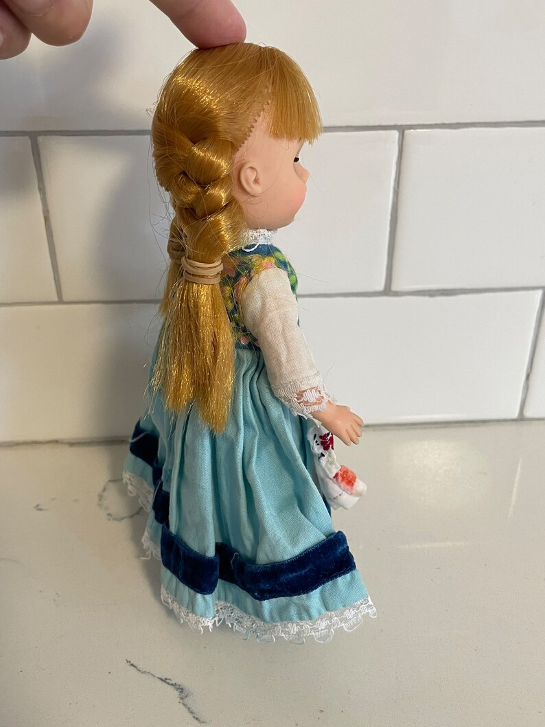 May include: A vintage porcelain doll with blonde hair in a braid, wearing a blue and white dress with a floral bodice and a blue velvet sash.
