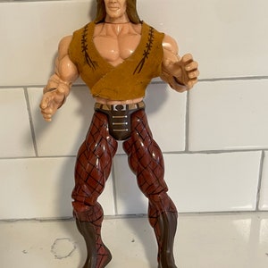 May include: A brown and red action figure with long brown hair, wearing a brown leather vest and brown pants with a patterned design.