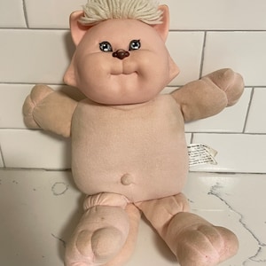 May include: A pink plush cat with a white yarn hairpiece. The cat has a brown nose and blue eyes. The cat is sitting on a white tiled surface.