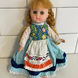 May include: A vintage porcelain doll with blonde hair in braids, wearing a blue and white dress with floral accents and a white apron. The doll has green eyes and a sweet expression.