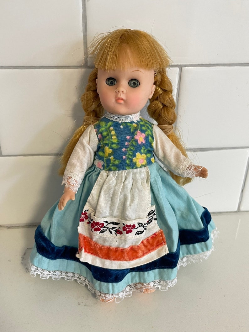May include: A vintage porcelain doll with blonde hair in braids, wearing a blue and white dress with floral accents and a white apron. The doll has green eyes and a sweet expression.