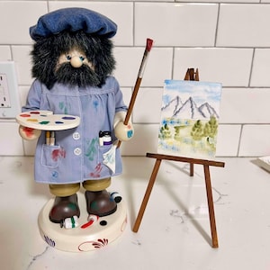 May include: A whimsical artist figurine with a blue beret and smock, holding a paintbrush and palette. A miniature painting on a wooden easel depicts a mountain landscape. The figurine stands on a decorative base with paint tubes.