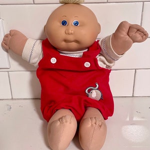 May include: A Cabbage Patch Kid doll with blonde hair, blue eyes, and a red and white outfit. The doll is wearing overalls with an elephant design.