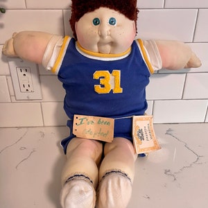 May include: A Cabbage Patch Kid doll wearing a blue and yellow basketball uniform with the number 31 on the front. The doll has brown hair, blue eyes, and a rosy complexion. The doll is sitting on a white tiled floor with a tag that says "I've been adopted!"