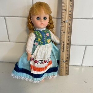 May include: A vintage doll with blonde hair, blue eyes, and a blue and white dress with a white apron. The doll is wearing a red and white embroidered belt and is holding a small white flower.
