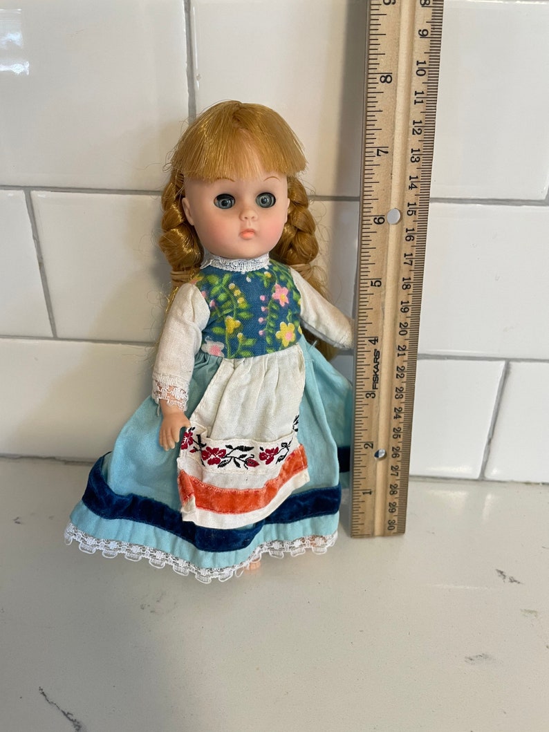 May include: A vintage doll with blonde hair, blue eyes, and a blue and white dress with a white apron. The doll is wearing a red and white embroidered belt and is holding a small white flower.