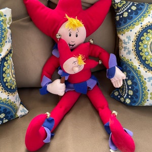 May include: A red and blue plush jester doll with a yellow jester hat and bells. The doll is holding a smaller red and blue plush jester doll with a yellow jester hat.