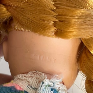 May include: A close-up of a porcelain doll's neck with the word "GINNY" printed on it. The doll has blonde hair and is wearing a blue and pink dress with white lace trim.