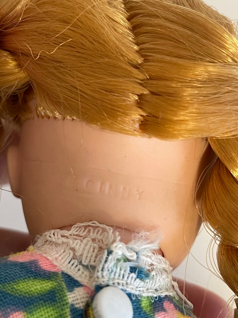 May include: A close-up of a porcelain doll's neck with the word "GINNY" printed on it. The doll has blonde hair and is wearing a blue and pink dress with white lace trim.