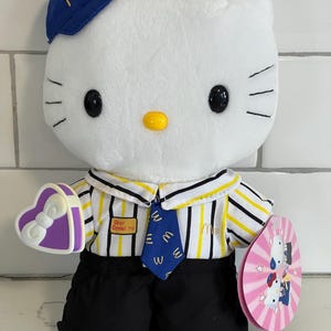 May include: White Hello Kitty plush toy wearing a blue and yellow McDonald's uniform with a purple heart-shaped box. The toy has a blue hat with the McDonald's 'M' logo and a yellow name tag that says 'Dear Daniel TM'.