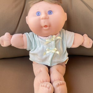 May include: A Cabbage Patch Kid doll with blonde hair, blue eyes, and a pink and blue outfit. The doll is sitting on a brown surface.