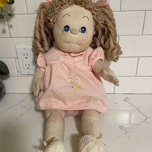May include: A vintage cloth doll with a hand-painted face, blue eyes, and freckles. The doll has curly blonde yarn hair tied with pink ribbons. It wears a pink dress with floral embroidery and white shoes.