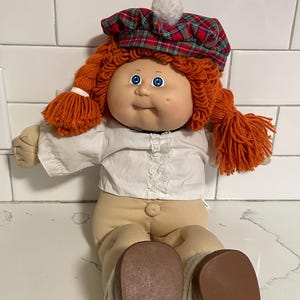 May include: A Cabbage Patch Kid doll with red yarn hair in pigtails, wearing a white shirt, tan pants, and a red and green plaid hat with a white pom-pom.