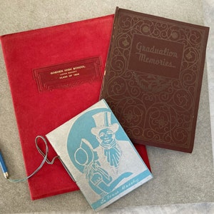 May include: A collection of vintage books and ephemera. Includes a red Goshen High School yearbook from 1936, a brown "Graduation Memories" book, and a small blue and white booklet with a gentleman illustration. A blue pencil rests nearby.