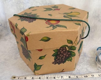 May include: Hexagonal gift box with a fruit pattern. The box is made of brown cardboard and features illustrations of grapes, peaches, and lemons. A green cord handle is attached to the top. A measuring tape is in the foreground.