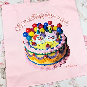 May include: Pink tote bag featuring a colorful cake with two clown faces, topped with balloons. The text "Clowning Around!" arches above the cake, with "P.S. Cakery" printed below. The bag has a simple, clean design.