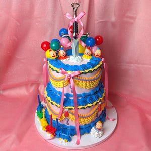 May include: A two-tiered cake with light purple frosting, decorated with blue and yellow ruffled piping, pink ribbons, and a silver sword topper. The cake features small clown figurines, colorful plastic spheres, and a toy train at the base.