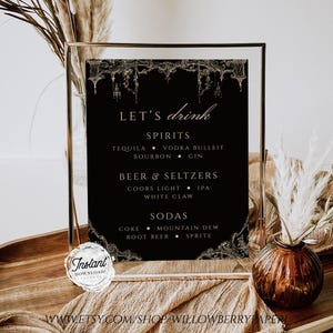 May include: A black and gold printable sign with the text "Let's drink" and a list of drinks including spirits, beer and seltzers, and sodas. The sign is framed in a gold frame and is sitting on a table with a brown vase and dried flowers.