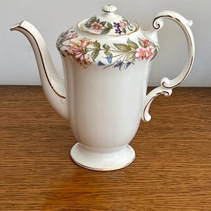 May include: A white porcelain coffee pot with a floral design. The pot has a curved spout, a decorative handle, and a lid with a floral pattern. Gold trim accents the edges of the pot and handle. The floral design includes pink, purple, and green flowers.