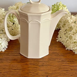 May include: A cream-colored ceramic teapot with an octagonal body, a matching lid with a loop handle, a curved spout, and a side handle. The upper portion of the teapot features a subtle embossed arch pattern along each of the eight flat sides.