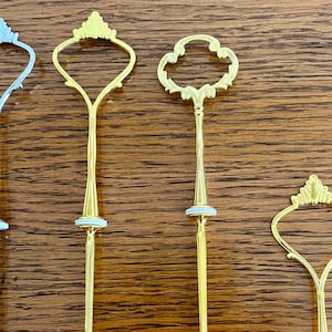 May include: Four cake decorating stands with different decorative tops. Two are gold, one is silver, and one is a simple gold loop.