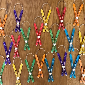 May include: A collection of 18 wooden ski ornaments, each painted in a different colour, including red, orange, yellow, green, blue, and purple. The skis are crossed and tied together with a white heart at the centre.