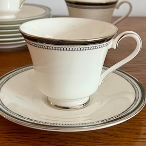 May include: White teacup and saucer set with a black chain-link design around the rim. The teacup has a curved handle and sits on a matching saucer. Additional teacups and saucers are visible in the background.