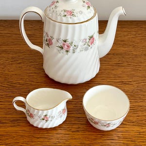 May include: A white porcelain tea set with a teapot, creamer, and teacup. The set features a floral pattern with pink roses and green leaves. The teapot has a lid and a curved handle. The creamer has a small handle and spout. The teacup is round.