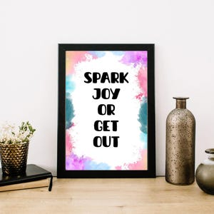 以下が含まれることがあります： 「SPARK JOY OR GET OUT」の黒い文字が入った額入りのプリント。背景はピンク、青、紫の色合いの水彩デザインです。フレームは黒で、プリントは木製の表面にあります。