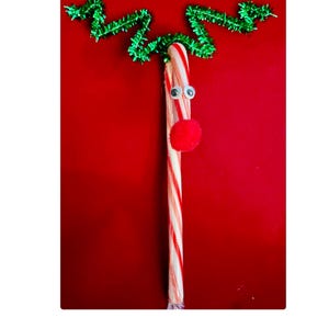 May include: A red and white candy cane with googly eyes and a red pom-pom nose, decorated with green pipe cleaners for antlers. The candy cane is on a red background.