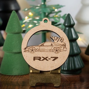 May include: A wooden Christmas ornament featuring a car design with the text "RX-7". The ornament is displayed on a small stand, surrounded by miniature green Christmas trees. The ornament is made of light brown wood.