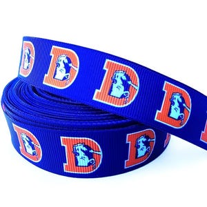 May include: A roll of blue ribbon featuring a repeating pattern of the letter "D" with a horse head logo in orange and light blue. The ribbon is coiled, with a portion unrolled, displaying the design.