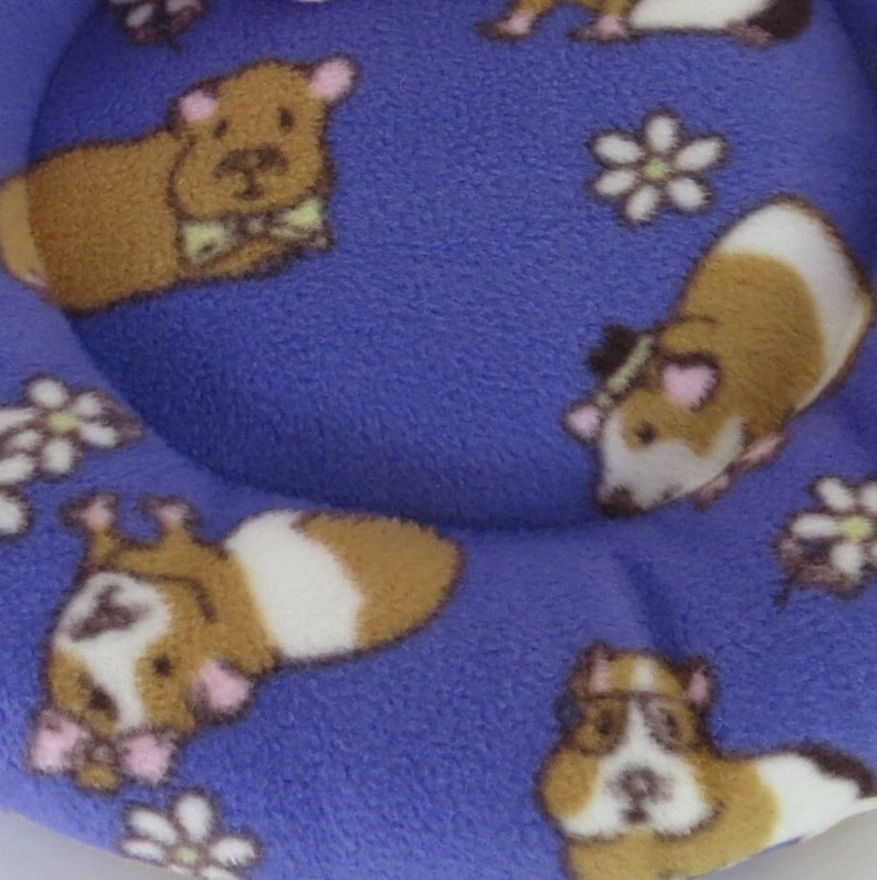 May include: A purple fleece pet bed with a brown and white guinea pig pattern. The bed is round and has a raised edge.
