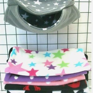 May include: A grey fleece pet hammock with white stars is suspended above a stack of colourful fleece pet bedding. The bedding displays various patterns, including stars, polka dots, and plaid, in colours such as pink, purple, and blue.