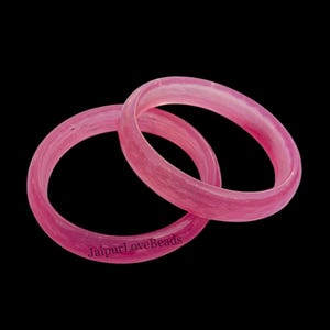 May include: Two pink bangles with a translucent appearance are displayed against a black background. The bangles are circular and appear to be made of a smooth material. The text "JaipurLoveBeads" is visible on one of the bangles.
