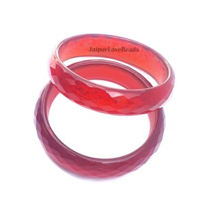 May include: Two faceted, red bangles are stacked on a white background. The bangles have a geometric design and a glossy finish. The text "JaipurLoveBeads" is visible on the top bangle.