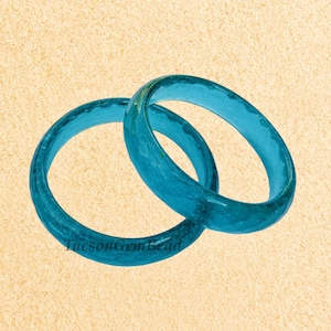 Op de afbeelding: Twee turquoise glazen armbanden, op elkaar gestapeld, tegen een lichtbruine achtergrond.