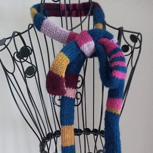 Könnte beinhalten: Ein handgestrickter Schal mit horizontalen Streifen in den Farben Dunkelblau, Senfgelb, Magenta und Hellrosa. Der Schal ist einfach geknotet und über eine schwarze Draht-Schneiderpuppe drapiert. Die Strickstruktur ist durchgehend gut erkennbar.