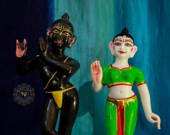 May include: Two hand-painted figurines of Hindu deities, one black and one white, standing on gold bases with floral designs. The black figurine is a male figure with a yellow sash and the white figurine is a female figure wearing a green and red outfit.