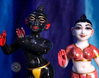 May include: Two statues featuring a dark-skinned figure with red accents and a light-skinned figure wearing a pink patterned top. The dark figure has a yellow loincloth, while the light figure has a matching pink skirt. Both have painted facial features.