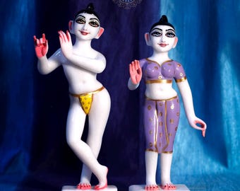 May include: Two white marble statues of figures standing on rectangular bases. The left figure wears a yellow loincloth with gold dots, while the right figure wears a purple top and skirt with gold accents. Both have painted faces and pink palms.