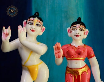 May include: Two white marble statues of deities, one male and one female, with painted details. The male statue is wearing a yellow dhoti and the female statue is wearing a red and gold sari.